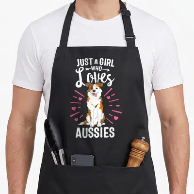 Aussy australian apron - Cartoon brown and white dog sitting with pink hearts and text — shown on a black cotton canvas apron against a white background