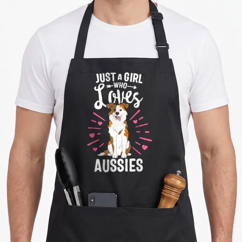Aussy australian apron - Cartoon brown and white dog sitting with pink hearts and text — shown on a black cotton canvas apron against a white background