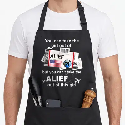 Proud boarding apron - Alief boarding pass graphic with barcode, American flag, and dotted airplane flight path — shown on a black cotton canvas apron against a white background