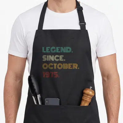 October 51st birthday apron - Stacked linear text reading LEGEND SINCE OCTOBER 1975 in teal beige yellow and orange-red — shown on a black cotton canvas apron against a white background