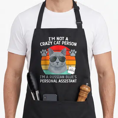 Blue cat apron - Grey Russian Blue cat with sunglasses, striped sunset, and personal assistant text — shown on a black cotton canvas apron against a white background