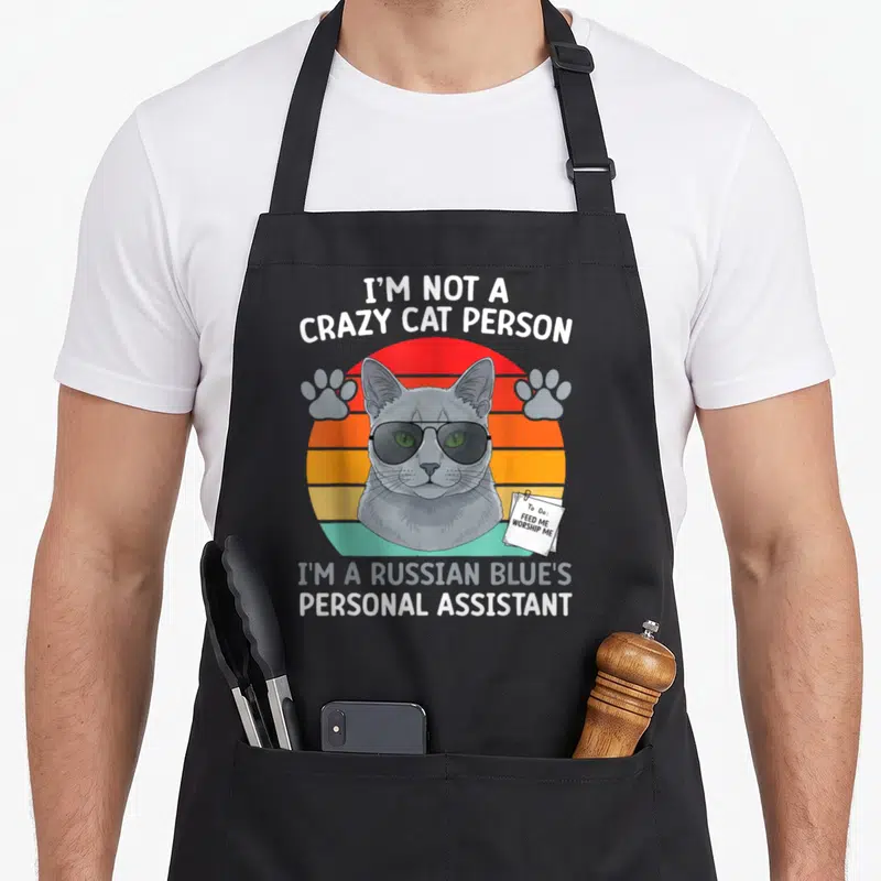 Blue cat apron - Grey Russian Blue cat with sunglasses, striped sunset, and personal assistant text — shown on a black cotton canvas apron against a white background