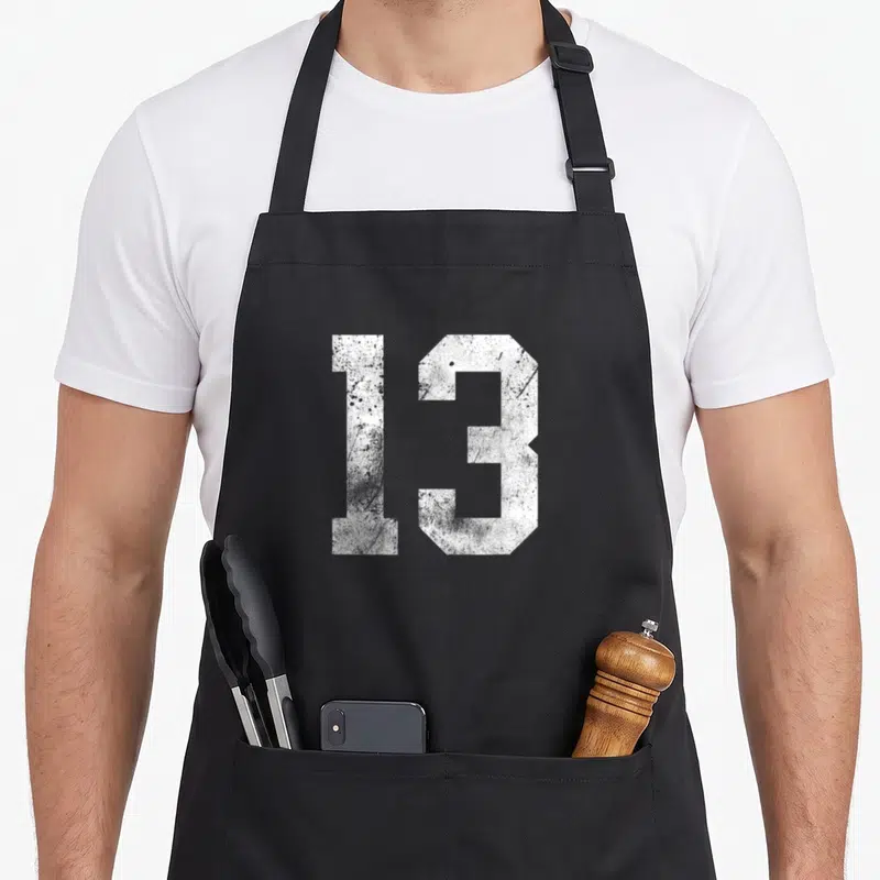 Distressed birthday apron - White number 13 in distressed block typography on grey background — shown on a black cotton canvas apron against a white background
