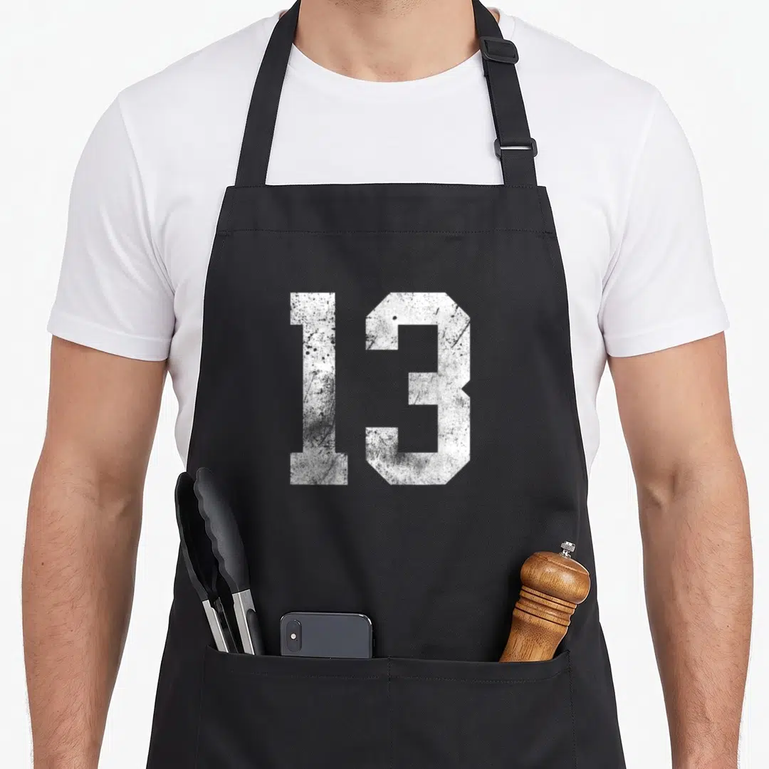 Distressed birthday apron - White number 13 in distressed block typography on grey background — shown on a black cotton canvas apron against a white background