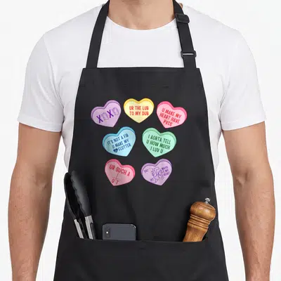 Cardiologist valentine apron - Seven colorful candy hearts with medical puns and anatomical drawings on black fabric — shown on a black cotton canvas apron against a white background