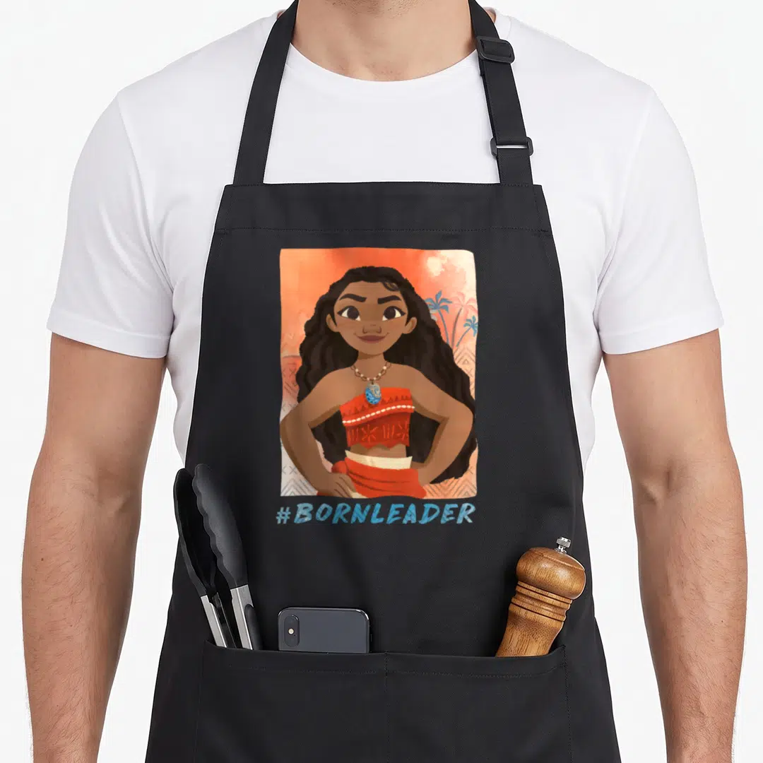 Bornleader novelty apron - Moana portrait with #BORNLEADER text on orange watercolor background with palm trees — shown on a black cotton canvas apron against a white background