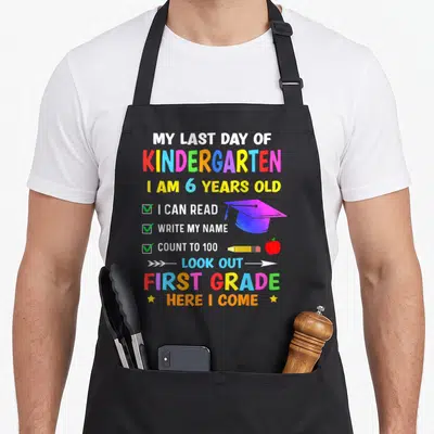 Kindergarten graduate apron - Colorful text listing kindergarten skills with graduation cap, pencil, apple, arrows — shown on a black cotton canvas apron against a white background