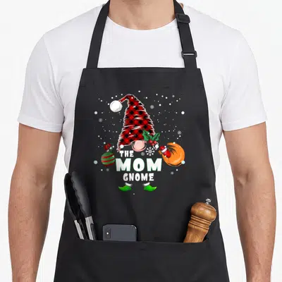 Cute gnome buffalo plaid christmas apron - THE MOM GNOME text with plaid hat, ornament, gift bag, and snowflakes — shown on a black cotton canvas apron against a white background