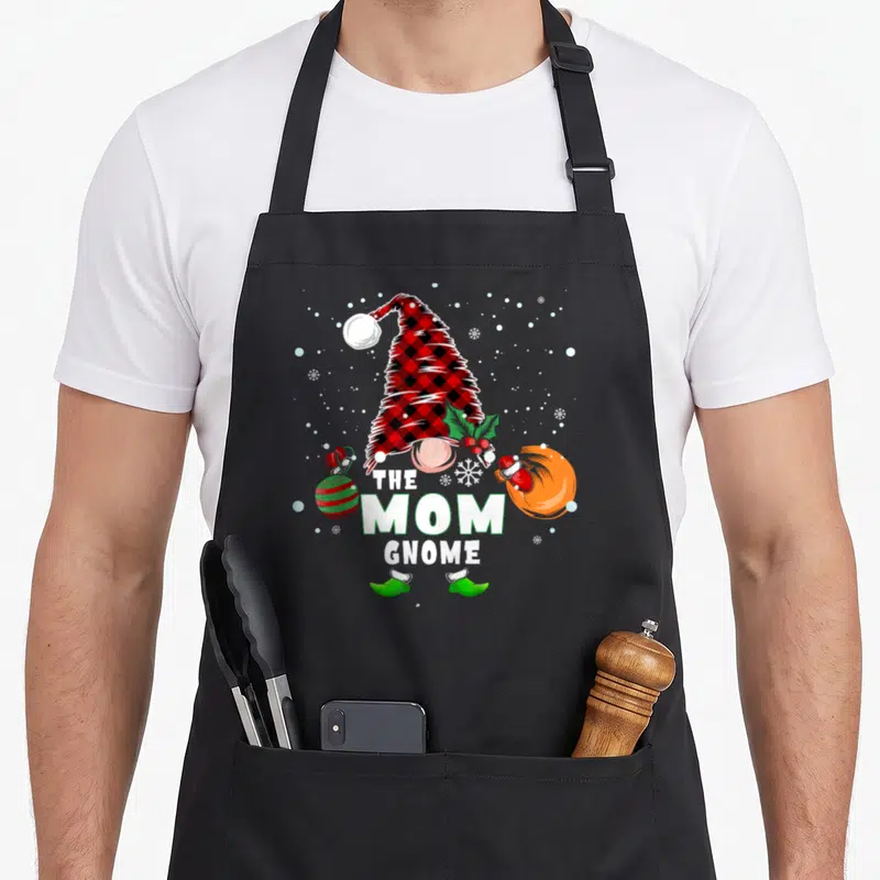 Cute gnome buffalo plaid christmas apron - THE MOM GNOME text with plaid hat, ornament, gift bag, and snowflakes — shown on a black cotton canvas apron against a white background