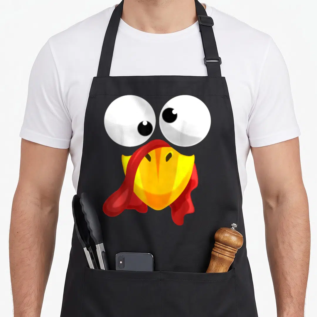 Clothe thanksgiving apron - Cartoon turkey face with big white eyes, yellow beak, and red wattle on black background — shown on a black cotton canvas apron against a white background