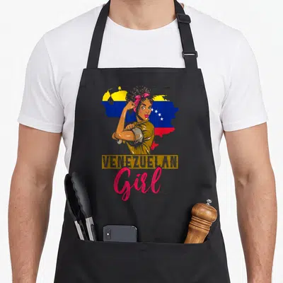 Proud venezuelan girl apron - Woman flexing arm over Venezuela map with VENEZUELAN Girl text in gold and pink — shown on a black cotton canvas apron against a white background