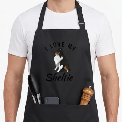 Sheltie adorable aprons - Cartoon tri-color Sheltie sitting between I Love My Sheltie text — shown on a black cotton canvas apron against a white background