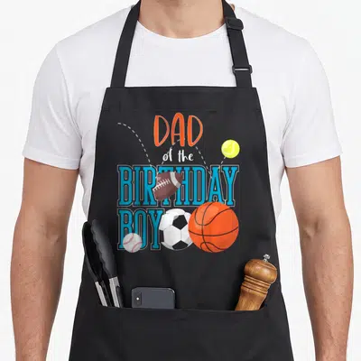 Sport family aprons - Dad of the Birthday Boy text with football, basketball, soccer, baseball, and tennis ball — shown on a black cotton canvas apron against a white background