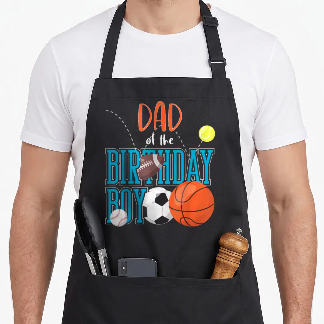 Sport family aprons - Dad of the Birthday Boy text with football, basketball, soccer, baseball, and tennis ball — shown on a black cotton canvas apron against a white background