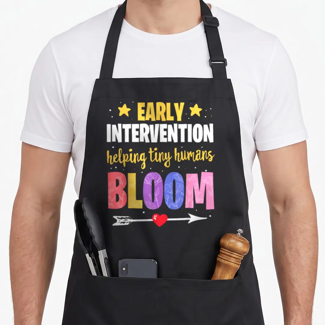Ecse teacher apron - Stacked text EARLY INTERVENTION helping tiny humans BLOOM with floral letters and heart arrow — shown on a black cotton canvas apron against a white background