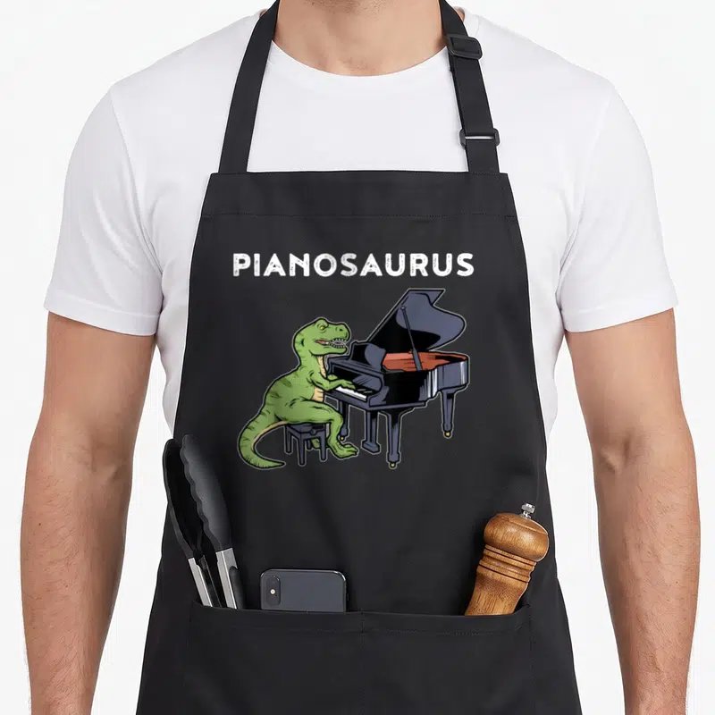 Grand piano apron - Green T-Rex playing black piano under white PIANOSAURUS text — shown on a black cotton canvas apron against a white background