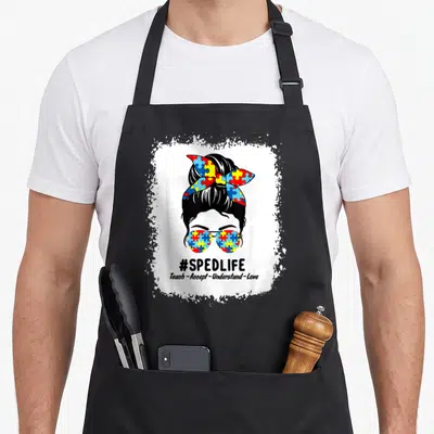 Special education apron - Cartoon woman with puzzle piece sunglasses and #SPEDLIFE text stacked below — shown on a black cotton canvas apron against a white background