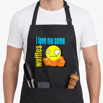 Scene waffles apron - Stacked text reading I love me some waffles with drooling emoji over waffle stack — shown on a black cotton canvas apron against a white background