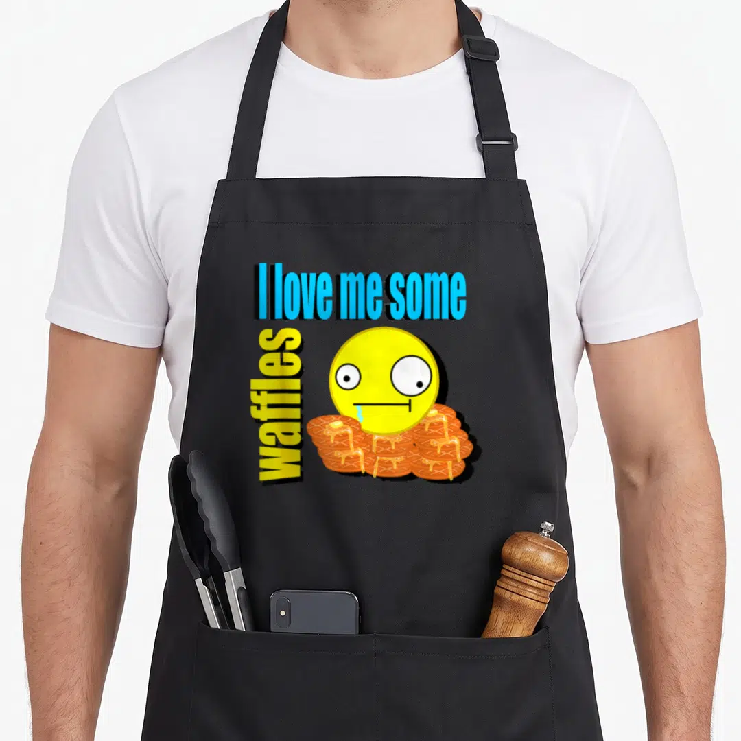 Scene waffles apron - Stacked text reading I love me some waffles with drooling emoji over waffle stack — shown on a black cotton canvas apron against a white background