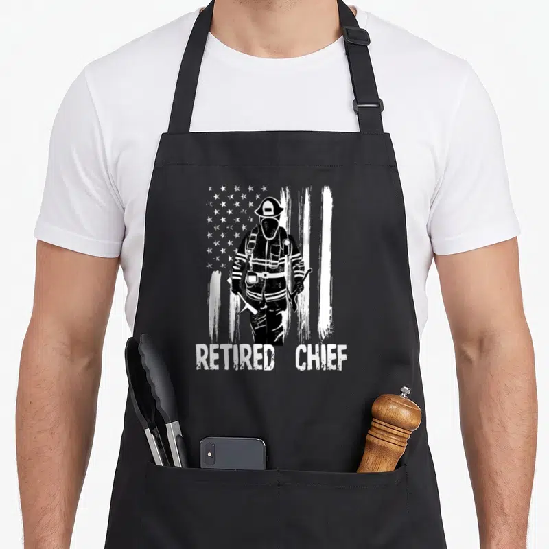 Chief firefighter cooking apron - Black firefighter silhouette holding axe in front of distressed white flag with text — shown on a black cotton canvas apron against a white background