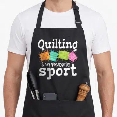 Handcrafting needlework apron - Quilting Is My Favorite Sport text with four colorful quilt squares and two hearts — shown on a black cotton canvas apron against a white background