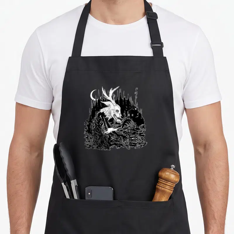 Wendigo horror apron - Black and white line art of a deer skull creature in a forest near a smoking cabin — shown on a black cotton canvas apron against a white background