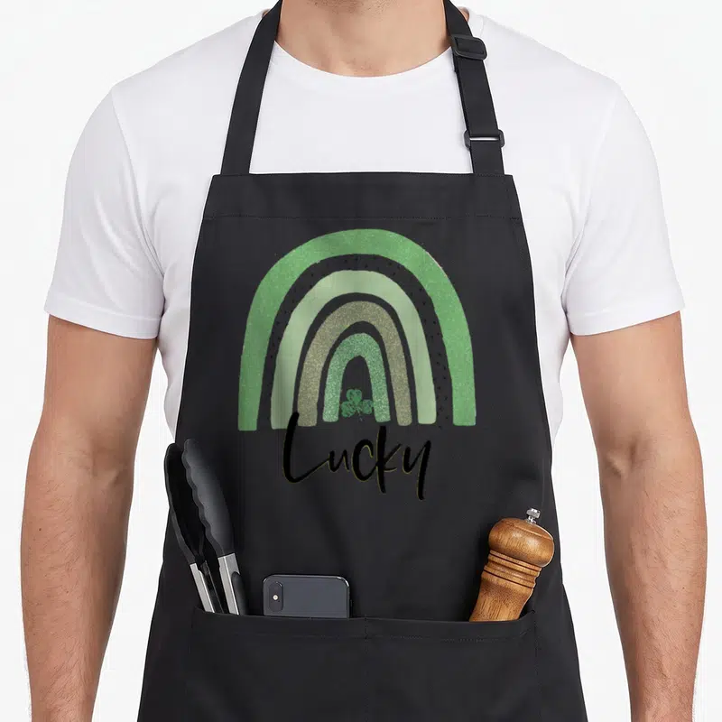 Lucky boho apron - Stylized rainbow in green grey gold black with lucky script and shamrock — shown on a black cotton canvas apron against a white background