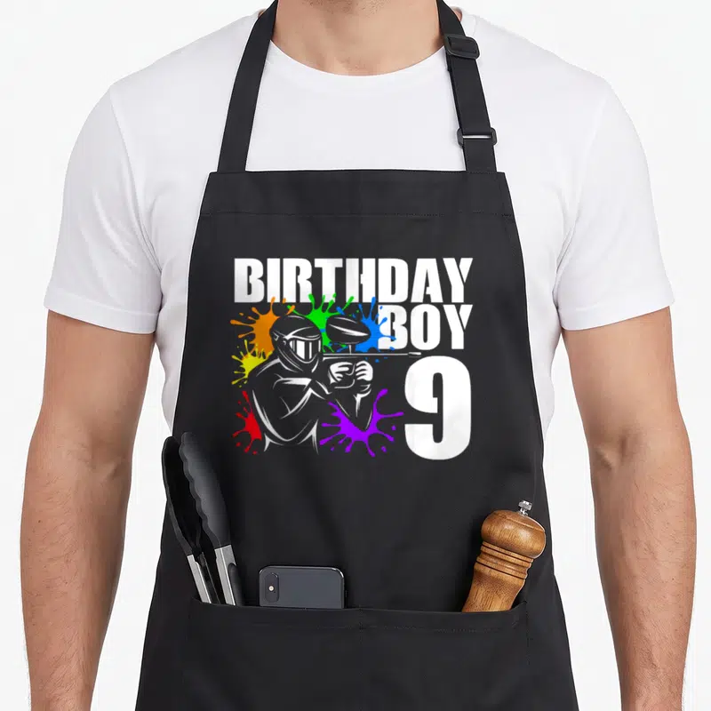 Paintball 9th birthday apron - Cartoon paintball player with BIRTHDAY BOY 9 text and colorful splats — shown on a black cotton canvas apron against a white background