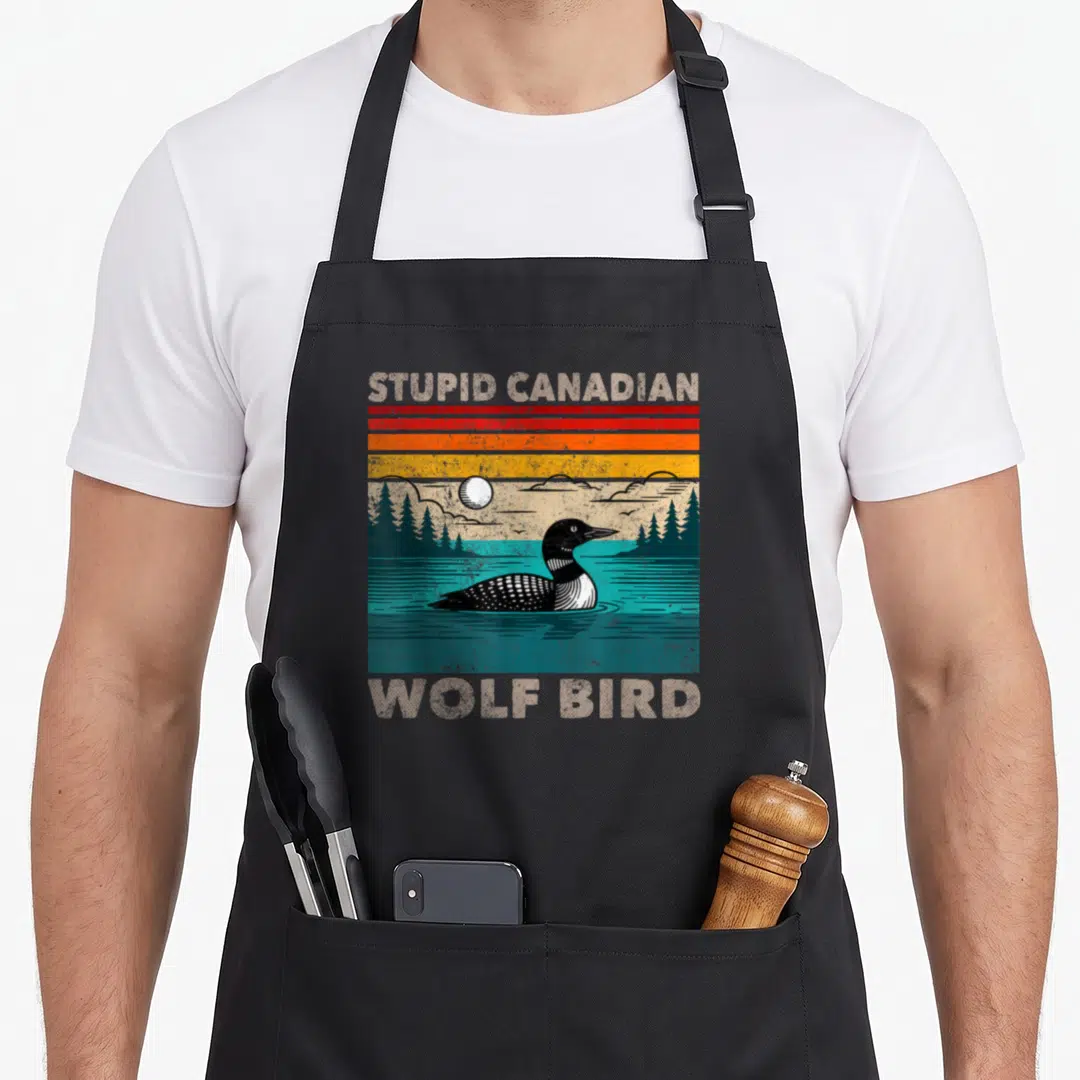 Stupid meme apron - Retro loon on teal water with STUPID CANADIAN WOLF BIRD text and striped sunset background — shown on a black cotton canvas apron against a white background