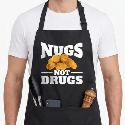 Nug drug chicken print apron - NUGS NOT DRUGS text above golden brown chicken nugget cluster with white lettering — shown on a black cotton canvas apron against a white background