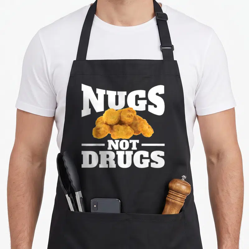 Nug drug chicken print apron - NUGS NOT DRUGS text above golden brown chicken nugget cluster with white lettering — shown on a black cotton canvas apron against a white background