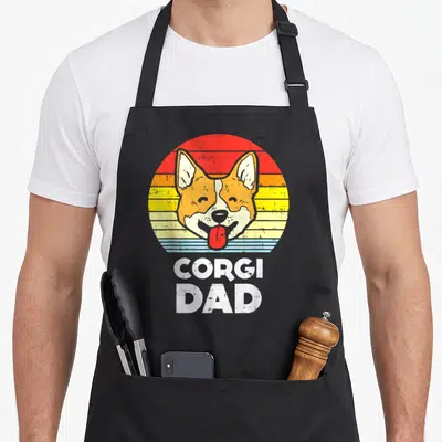 Sunset welsh apron - Retro corgi head on orange red yellow striped circle with white CORGI DAD text below — shown on a black cotton canvas apron against a white background