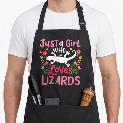 Funny lizard apron - White lizard silhouette with pink text and floral wreath in pink green orange — shown on a black cotton canvas apron against a white background