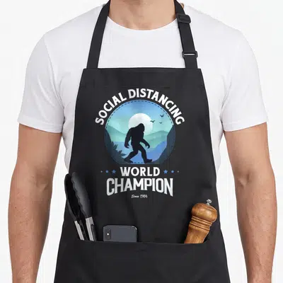 Distance bigfoot apron - Bigfoot silhouette in blue landscape with SOCIAL DISTANCING WORLD CHAMPION text — shown on a black cotton canvas apron against a white background