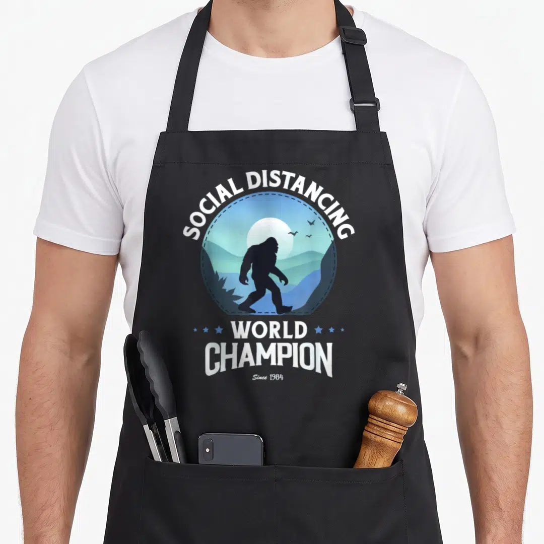 Distance bigfoot apron - Bigfoot silhouette in blue landscape with SOCIAL DISTANCING WORLD CHAMPION text — shown on a black cotton canvas apron against a white background