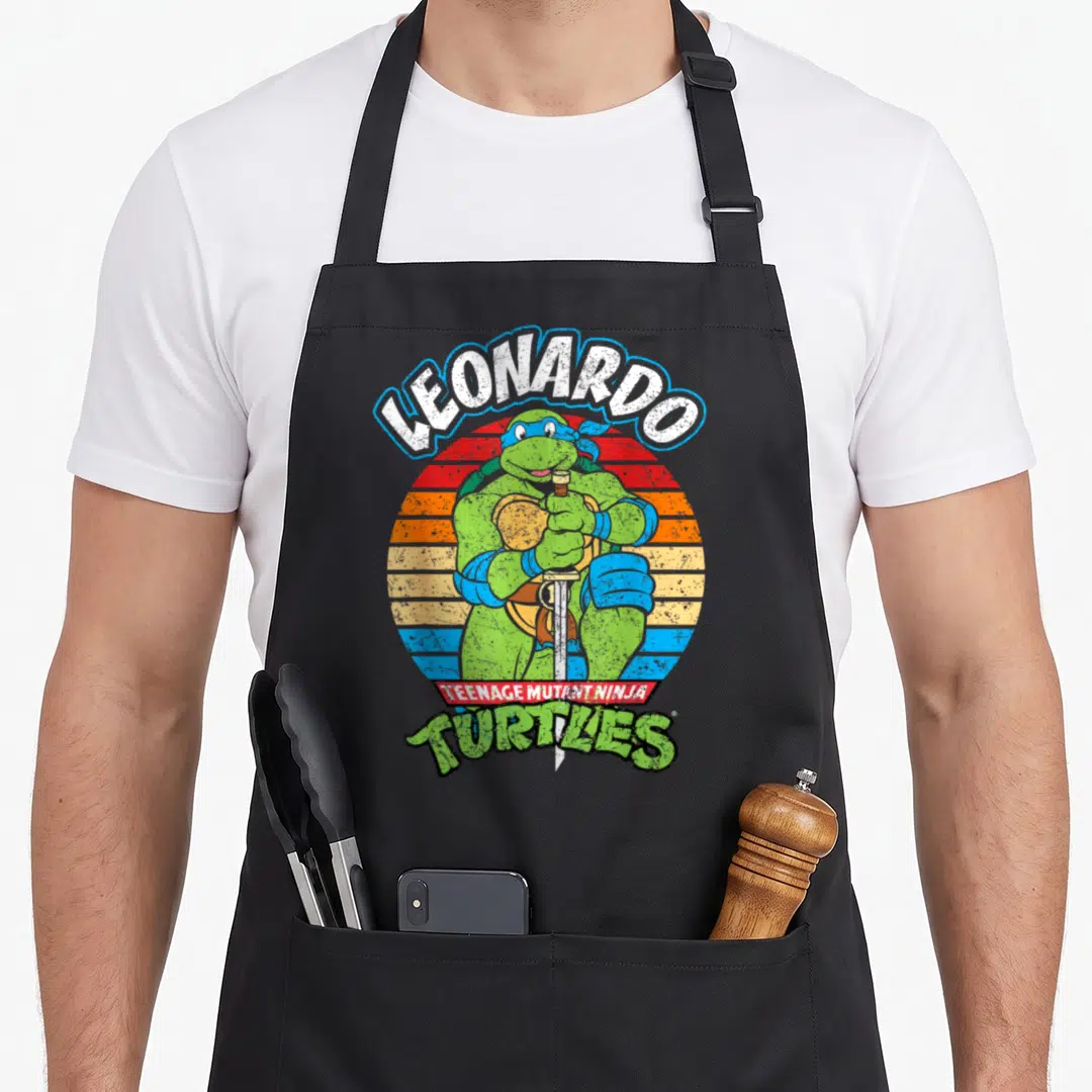 Ready teenage cooking aprons - Leonardo holding katanas in front of a striped retro sun with arched text — shown on a black cotton canvas apron against a white background
