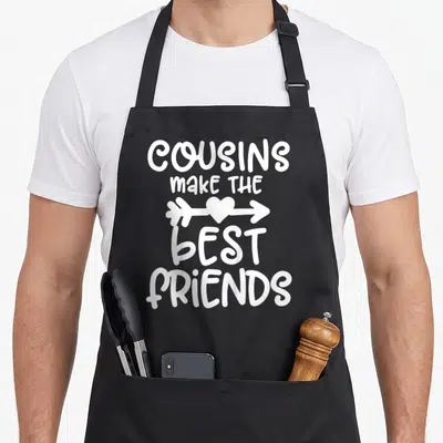 Toddler friends apron - White text COUSINS MAKE THE BEST FRIENDS with heart and arrow on black background — shown on a black cotton canvas apron against a white background
