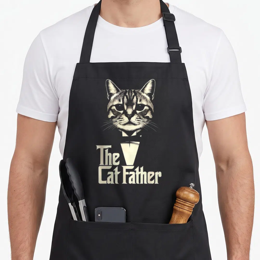 Catfather cat dad apron - Line art tabby cat head above stacked The Cat Father text separated by a tuxedo collar graphic — shown on a black cotton canvas apron against a white background