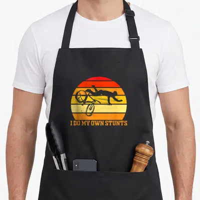 Cycle cycling apron - Grey silhouette of person falling off bike against red orange yellow striped sun with text I DO — shown on a black cotton canvas apron against a white background