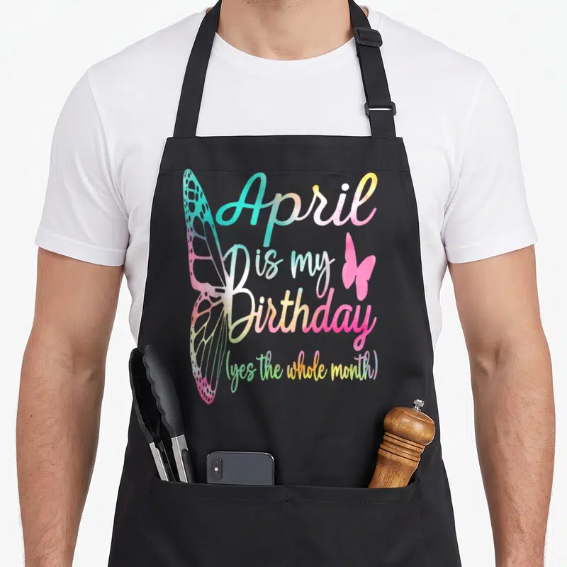 Funny birthday april apron - April is my Birthday text with large and small pink butterflies in pastel gradient colors — shown on a black cotton canvas apron against a white background