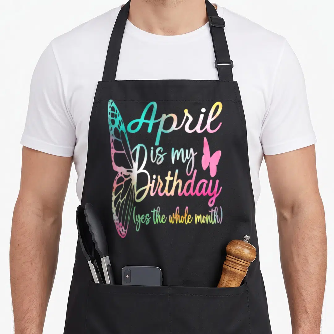 Funny birthday april apron - April is my Birthday text with large and small pink butterflies in pastel gradient colors — shown on a black cotton canvas apron against a white background