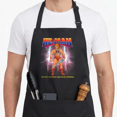 Most universe apron - He-Man holding a sword with lightning bolts and the Most Powerful Man slogan — shown on a black cotton canvas apron against a white background