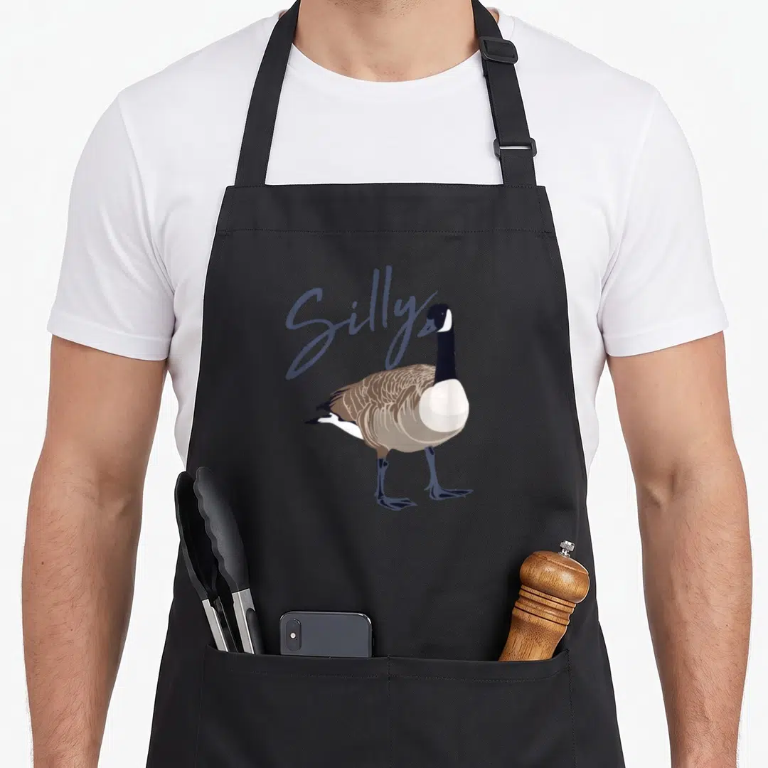 Canadian silly apron - Cartoon goose standing with Silly script in blue-grey behind it — shown on a black cotton canvas apron against a white background