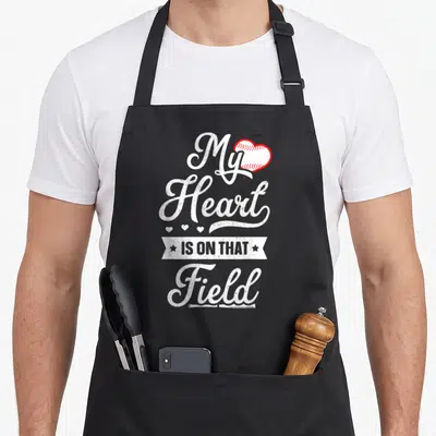 Funny field baseball apron - Distressed white text My Heart IS ON THAT Field with red stitched heart and small stars — shown on a black cotton canvas apron against a white background