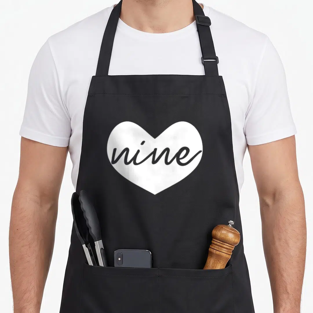 Ninth chef outfit apron - White heart shape containing the word nine in grey cursive script — shown on a black cotton canvas apron against a white background