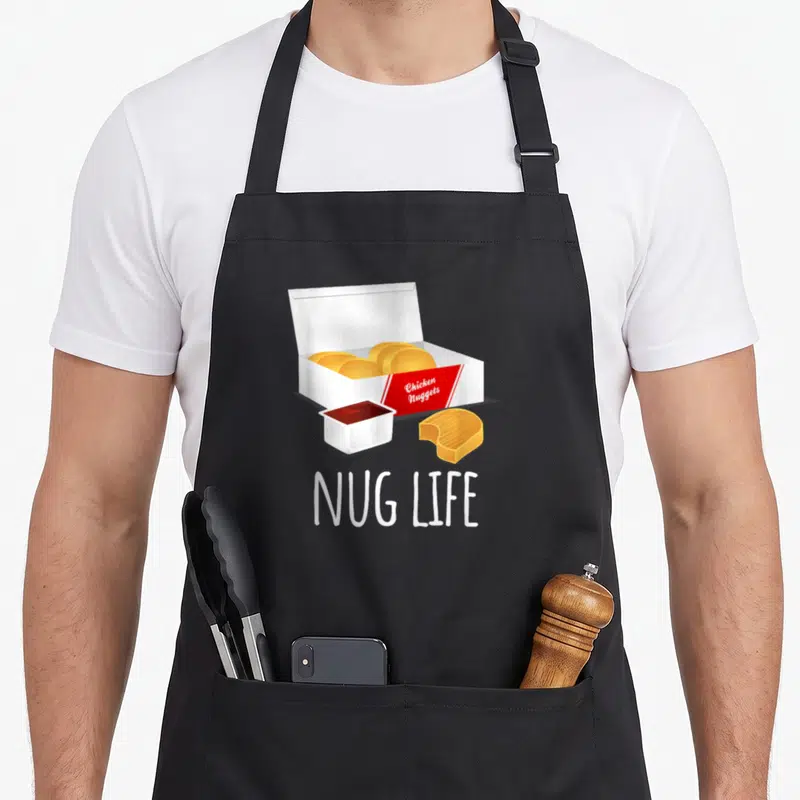 Nug nugget chicken print apron - Cartoon open box of golden chicken nuggets with red label, sauce cup, bitten nugget — shown on a black cotton canvas apron against a white background
