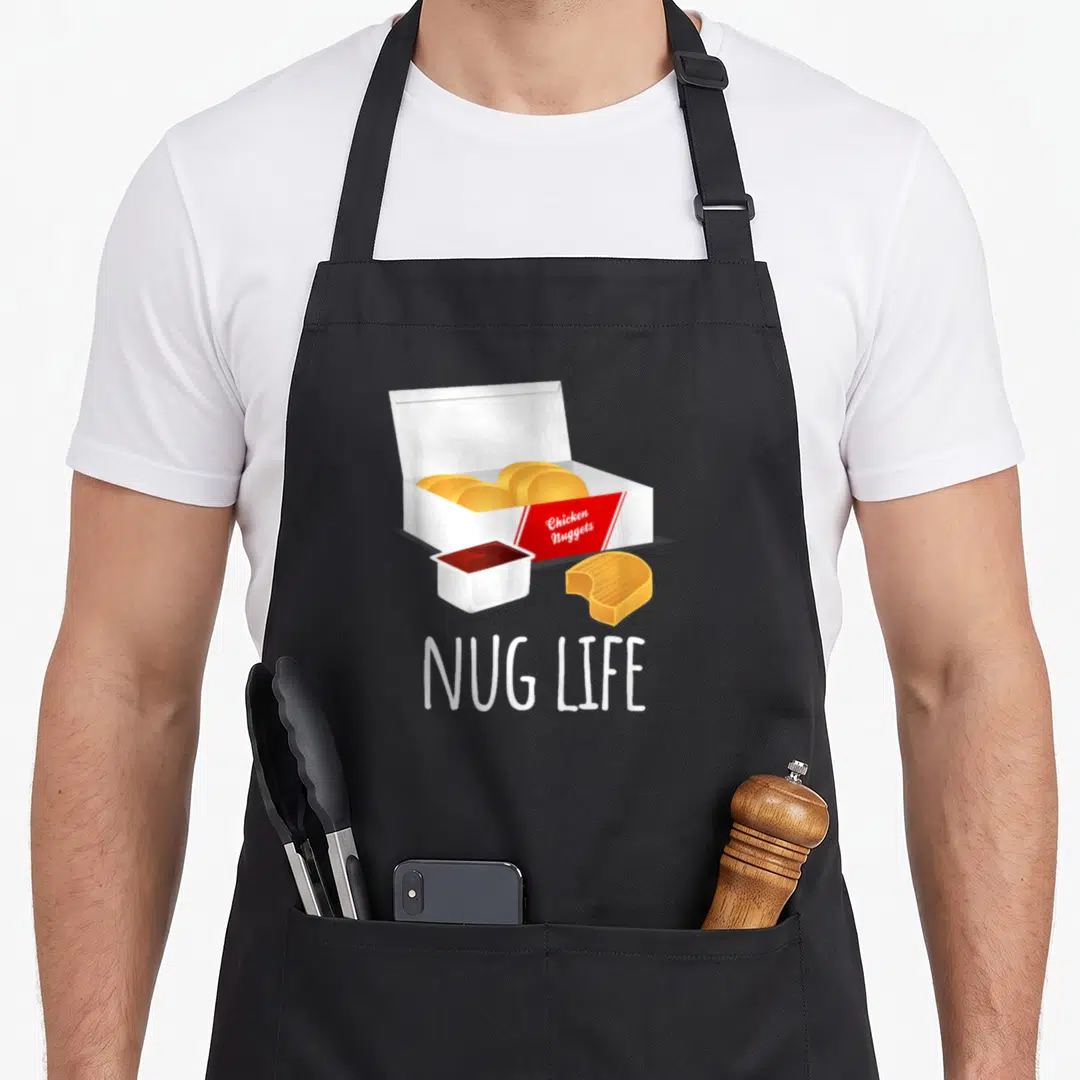 Nug nugget chicken print apron - Cartoon open box of golden chicken nuggets with red label, sauce cup, bitten nugget — shown on a black cotton canvas apron against a white background