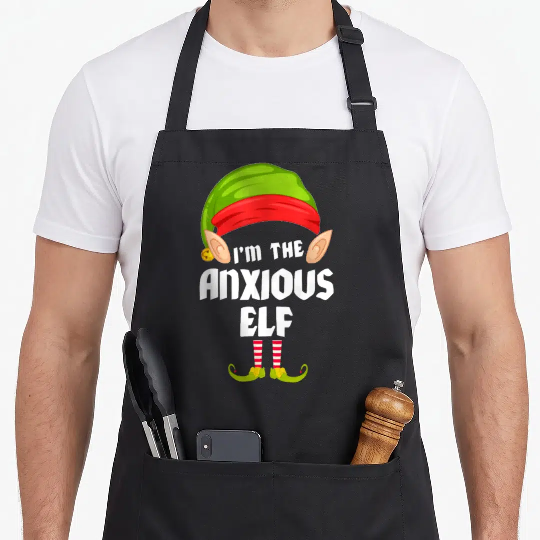 Anxious christmas elf apron - I'M THE ANXIOUS ELF text surrounded by elf hat, ears, striped socks and yellow shoes — shown on a black cotton canvas apron against a white background