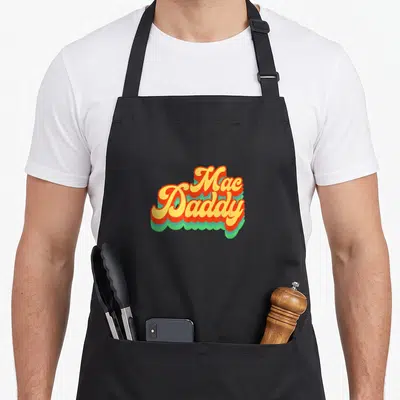 Mac daddy apron - Stacked Mac Daddy cursive text in yellow red orange and green retro typography design — shown on a black cotton canvas apron against a white background