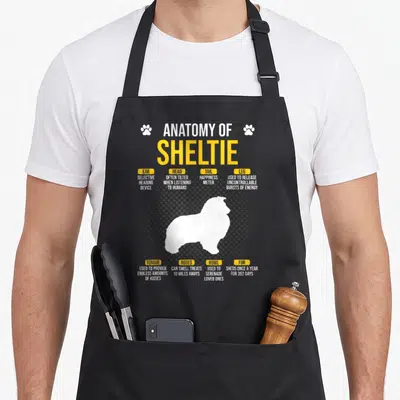 Anatomy sheltie dog apron - White dog silhouette on halftone dots with yellow labels describing anatomy like selective — shown on a black cotton canvas apron against a white background